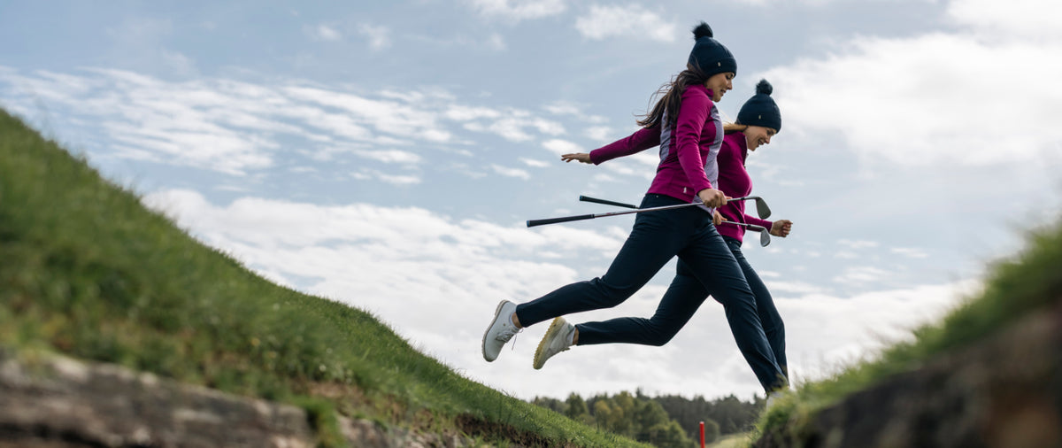 Two women jumping over a waterway at a golf course wearing Glenmuir navy bobble hats with thermal lining, Glenmuir ladies navy trousers and Glenmuir women's mid layer jersey jacket in raspberry and print and raspberry quarter zip sweater.