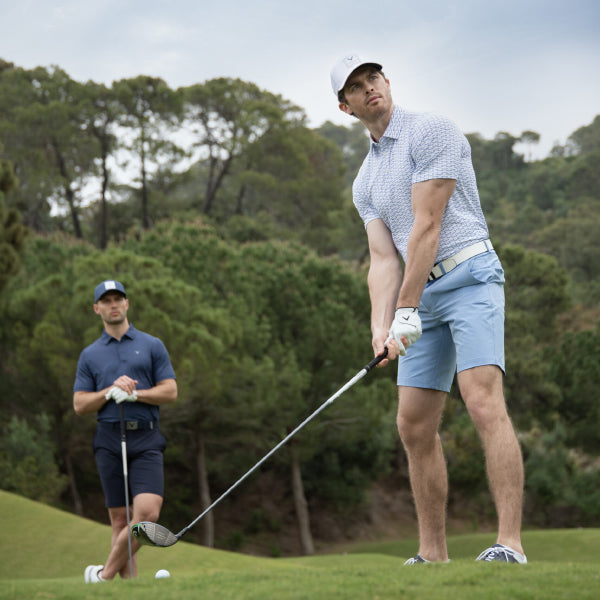A male golfer in a light blue print polo shirt, light blue shorts, and a white cap from Callaway golf, is lining up his shot on a golf course, with another man in a Callaway navy polo and shorts standing in the background.