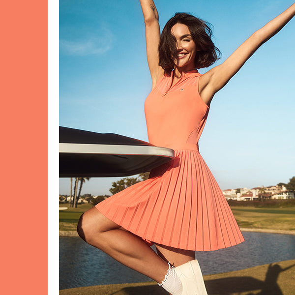 Woman golfer on golf buggy in Original Penguin pleated sleeveless coral golf dress available at GolfGarb