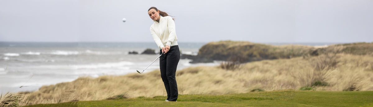 Female golfer wearing Ping thermal black trousers and a cream mid layer chipping a shot on a links golf course
