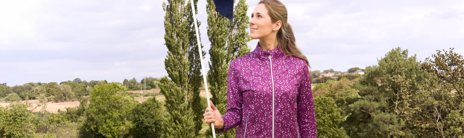 A woman wearing a Pure Golf black forest grove morello orchard print full zip-up mid layer jacket standing on the golf course holding a golf flagpole, with trees and a cloudy sky in the background.