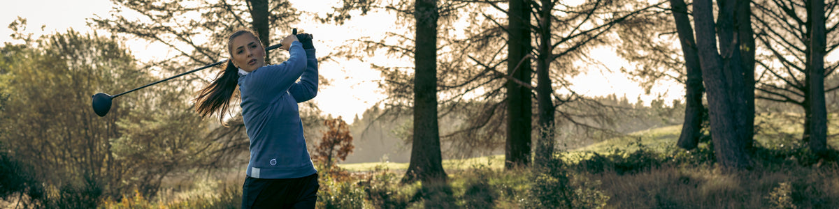 Female golfer completing her shot with a driver wearing the Sunderland of Scotland quarter zip in mineral blue. a wooded area is shown behind her.