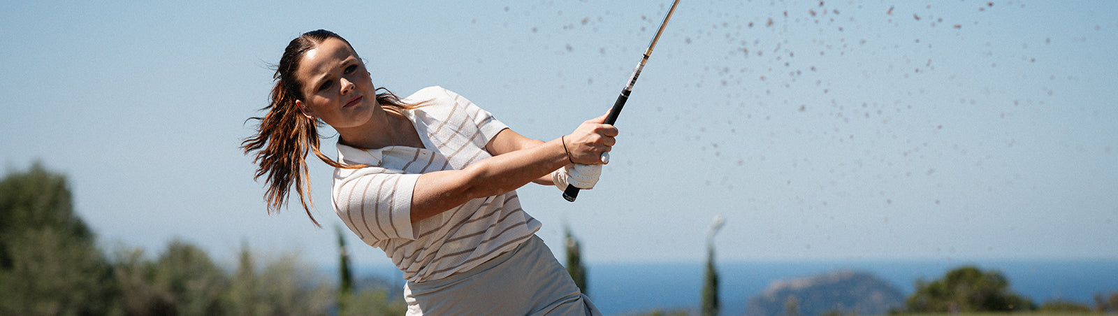 Female golfer completing her shot with bunker sand flying around her club wearing the adidas mesh knit short sleeve polo in white and beige stripe.